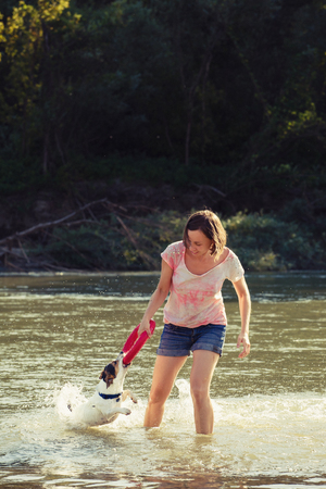 Young cheerful woman holding toy and playing with dog in water of river.の写真素材