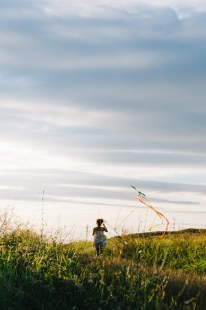 Back view of girl holding kite and running in field under sky with overcast.の写真素材