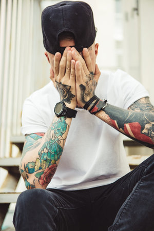 Young tattooed man in cap covering face with hands while posing on stairs.
