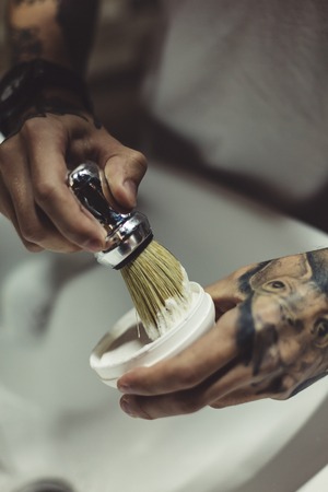 Crop shot in close-up of tattooed stylist mixing foam with shaving brush.の写真素材