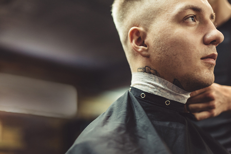 Crop stylist preparing young tattooed man in chair for shaving in barbershop.の写真素材