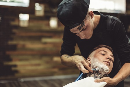 Crop barber using razor and shaving tattooed man in chair.の写真素材