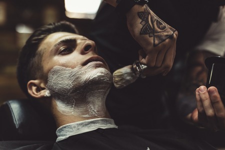 Crop stylish applying foam on customers cheeks for shaving while working in barbershop.の写真素材