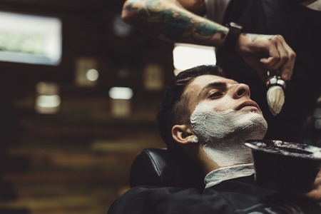 Crop stylish applying foam on customers cheeks for shaving while working in barbershop.の写真素材