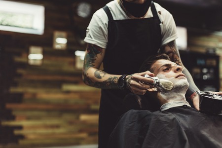 Crop stylish applying foam on customers cheeks for shaving while working in barbershop.の写真素材