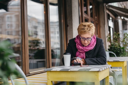 Adult blonde woman sitting in outside cafe and browsing smartphone while having coffee.の写真素材