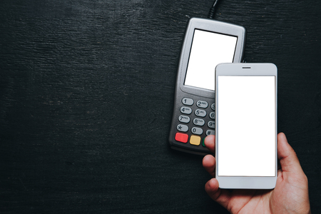 Top view of person using contactless payment with smartphone. Clear white mockup screen template of phone and pos terminal on dark wooden desk with side copy spaceの写真素材