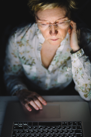 Middle-aged woman working on laptop in dark roomの写真素材