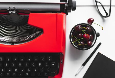 Crop view of white desk workplace with bright red vintage typewriter and little cup with cherryの写真素材