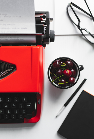Crop view of white desk workplace with bright red vintage typewriter and little cup with cherryの写真素材