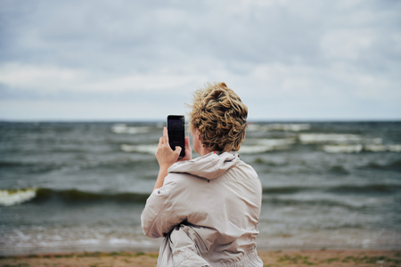 Back view of adult woman in warm jacket using smartphone to take picture of beautiful waving sea on cloudy dayの写真素材