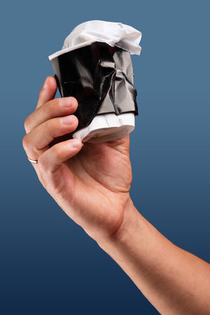 Male hand holds, raised black crumpled paper cup of tea or coffee with lid.