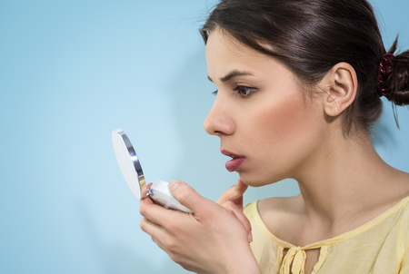 Woman with opened mirror, looking on her face on blue background.の写真素材