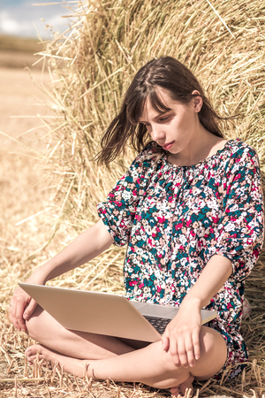 the brunette girl sitting cross-legged  on the hay and working with computer. beautiful smileの写真素材