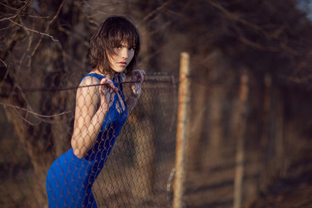 Beautiful girl in evening blue dress stands and looks over the fence の写真素材