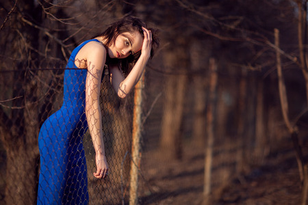 Beautiful girl in evening blue dress stands and looks over the fence の写真素材