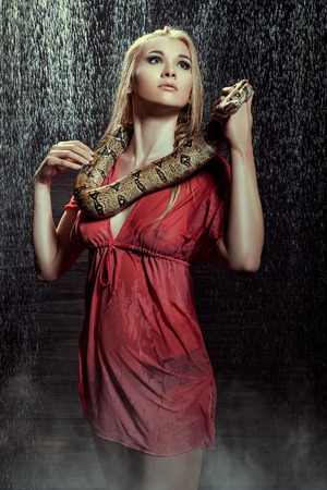 Beautiful blonde girl in a red negligee standing with wet snake in the rain の写真素材