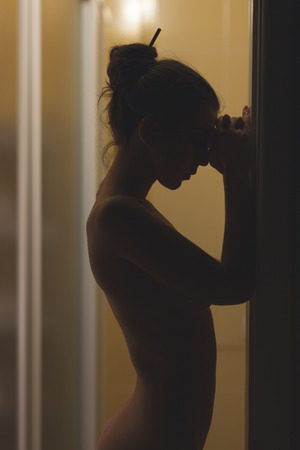 Naked girl standing sideways, his head leaning on a wall in a dark room.の写真素材