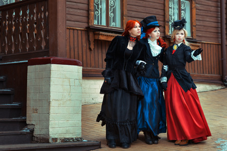 Three girls in retro dress standing and looking around the house on the street.の写真素材