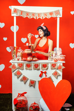 Pin-up girl style girl in the booth, she looks at the cake, near a lot of sweets.の写真素材