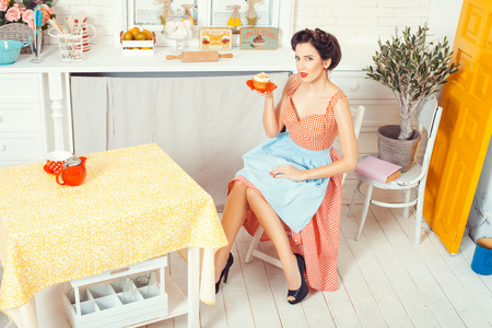 Pin-up girl style. The view from the top. Woman in an apron sitting on a chair in the middle of the kitchen and holding a saucer with muffin.の写真素材
