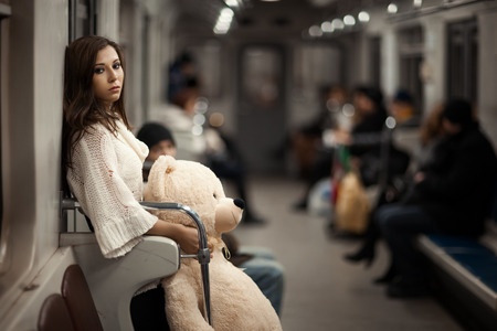 Sad girl with toy bear in her hands, she in a subway car. Rear people but they are very vague and can not be found. Photo toned.の写真素材