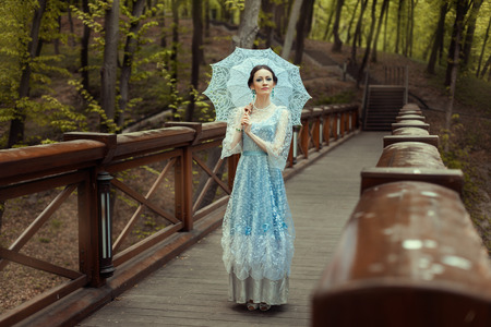 Woman standing on the bridge in a fantastic magical forest. She is dressed in a vintage blue dress. In the hands holding an oldfashioned knitted umbrella.の写真素材