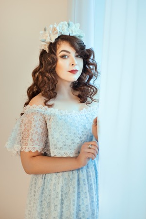 Beautiful young woman standing by a window holding hands curtain. Soft focus with small depth of field.の写真素材
