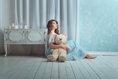 Beautiful woman with overweight sitting on the floor in the room. In her hands she holds toy bear.の写真素材