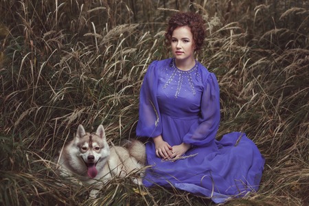 The girl sits in a high grass, her dog huskies lies nearby.の写真素材
