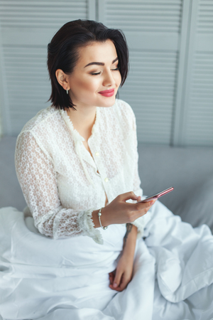 Pretty woman smiling in the morning reading the messages on the phone, she sits on the bed.の写真素材