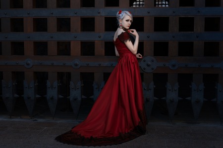 Woman in a long red dress, she is near the gate of an ancient castle.の写真素材