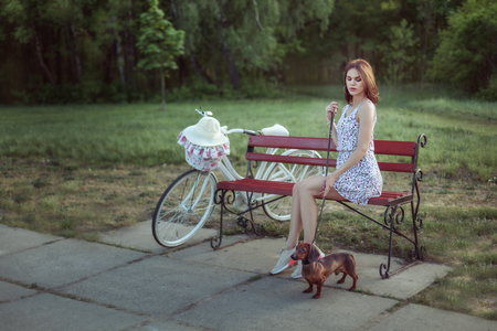 Young woman is sitting in the park on the bench. Nearby is her bicycle, a woman is holding a dachshund on a leash.の写真素材