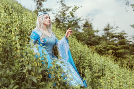Beautiful elf woman in a blue dress is sitting in the grass on a hill.の写真素材