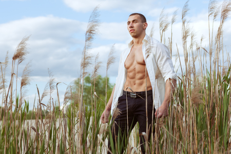 Young macho man in an unbuttoned white shirt standing in grass.の写真素材