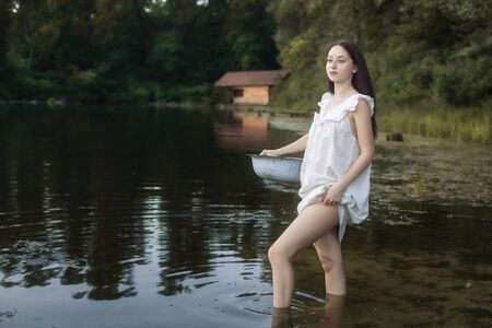 Young laundress stands in a river and holds a basin in her hands. She is preparing to wash clothes.の写真素材