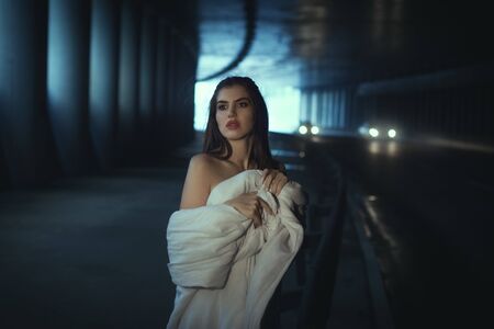 Young woman standing near a road in a tunnel, she is wrapped in a white blanket.の写真素材