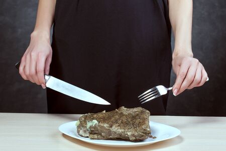Woman with beautiful hands holds a fork and knife. In front of her is a plate with a piece of meat.の写真素材