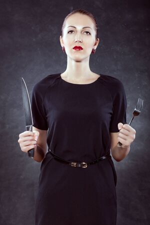 Portrait of a beautiful young woman on a black background with a knife and a fork in her hands.の写真素材