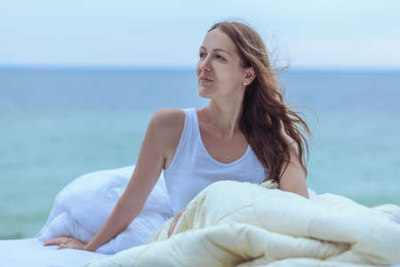 The bed is on the ocean. On the bed is a beautiful woman, in her hands a bouquet of flowers. The woman on the bed looks at the ocean.の写真素材