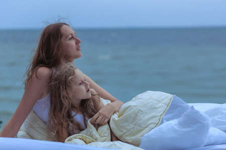 Woman and child look at the ocean while sitting in bed. They woke up on the ocean shore and dream of enjoying a beautiful landscape.の写真素材
