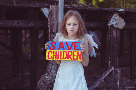 A girl with a poster in her hands asks to protect children. It is against the backdrop of a burned-out house from the war.の写真素材