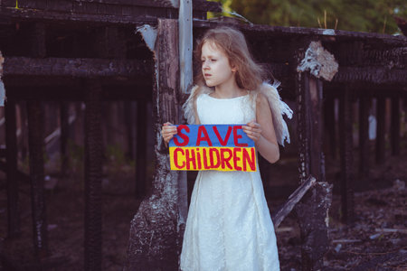 A girl with a poster in her hands asks to protect children. It is against the backdrop of a burned-out house from the war.の写真素材