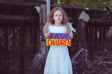 A girl with a poster in her hands asks to protect children. It is against the backdrop of a burned-out house from the war.の写真素材