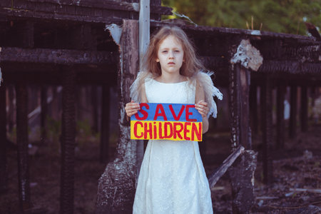 A girl with a poster in her hands asks to protect children. It is against the backdrop of a burned-out house from the war.の写真素材