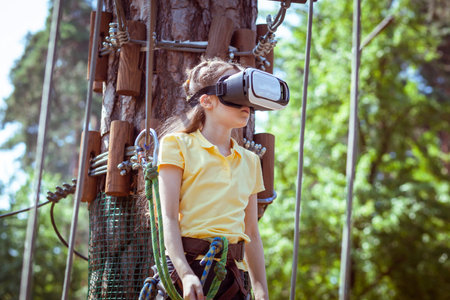 The girl plays a virtual game, she goes in for sports virtually. Virtual reality glasses help in sports. Virtual glasses are immersed in a children's forest rope adventure park.の写真素材