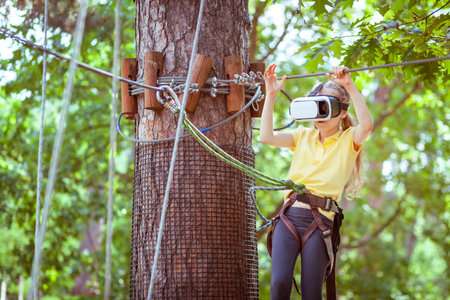 The girl plays a virtual game, she goes in for sports virtually. Virtual reality glasses help in sports. Virtual glasses are immersed in a children's forest rope adventure park.の写真素材