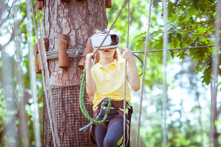 The girl plays a virtual game, she goes in for sports virtually. Virtual reality glasses help in sports. Virtual glasses are immersed in a children's forest rope adventure park.の写真素材