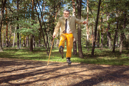 Funny old man is roller skating in the park. An active grandfather with a walking stick dances and makes faces. Grandfather with a white beard.の写真素材