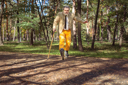 Funny old man is roller skating in the park. An active grandfather with a walking stick dances and makes faces. Grandfather with a white beard.の写真素材